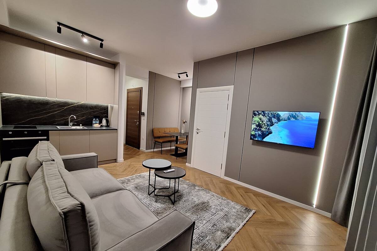 A modern living room with a sofa, a small table, a TV mounted on the wall, and a kitchen area with a sink and cabinets.
