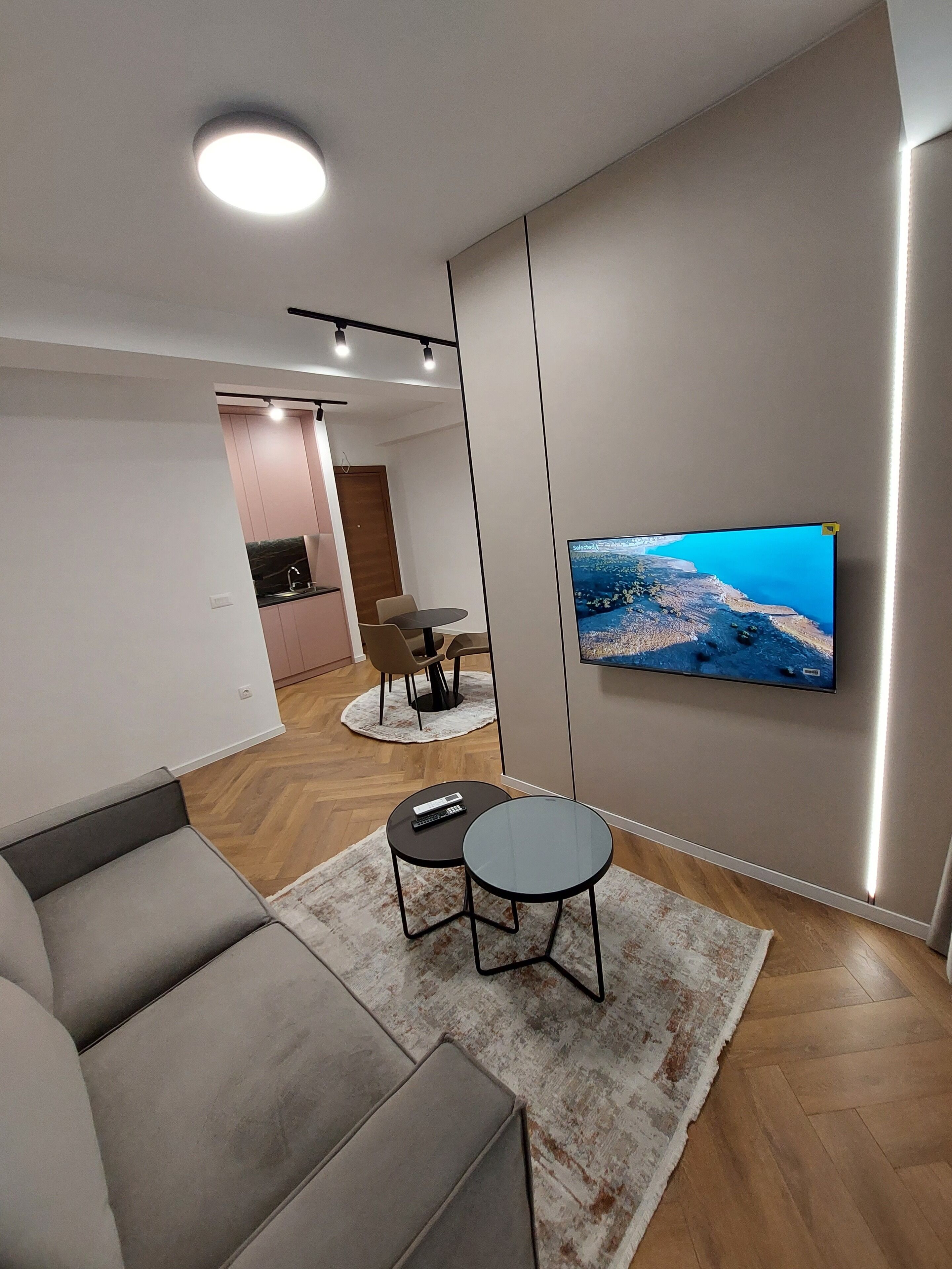 Ett modernt vardagsrum med en grå soffa, två små bord, en tv på väggen och ett kök i bakgrunden.