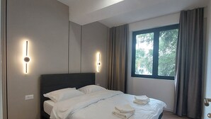 A modern bedroom with a large bed, two wall-mounted light fixtures, a window with curtains, and a wooden floor.