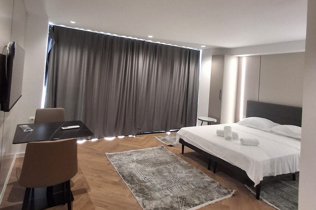 A modern hotel room with a bed, a desk, a chair, a TV, and a large window with curtains.