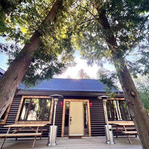 Una cabaña con fachada de madera, ventanas amplias y una puerta. Hay dos zonas de estar al aire libre con bancos y mesas.