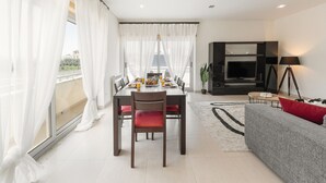 A modern living room with a dining area, a sofa, a TV, and a balcony with curtains. - Nazaré Beach House - 5 minutes walking to the beach (Leiria)