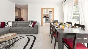 A modern living room with a grey sectional sofa, a dining table with red chairs, and a kitchen in the background. - Nazaré Beach House - 5 minutes walking to the beach (Leiria)