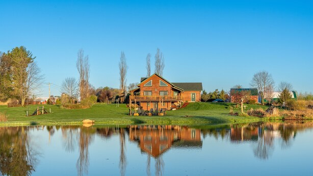 A large wooden cabin with a balcony, situated next to a calm lake with reflections. - Luxe Amish Log Cabin | 10 Acres | Private Pond | Kayak | Pizza Oven | ATV Trails (Stratford)