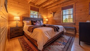 A wooden cabin bedroom with a bed, two bedside lamps, a rug, and a wooden dresser. - Luxe Amish Log Cabin | 10 Acres | Private Pond | Kayak | Pizza Oven | ATV Trails (Stratford)