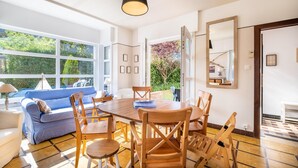 A modern dining area with a wooden table and chairs, a blue sofa, and large windows offering a view of greenery. - Apartment in Koksijde With Terrace & Garden (Koksijde)
