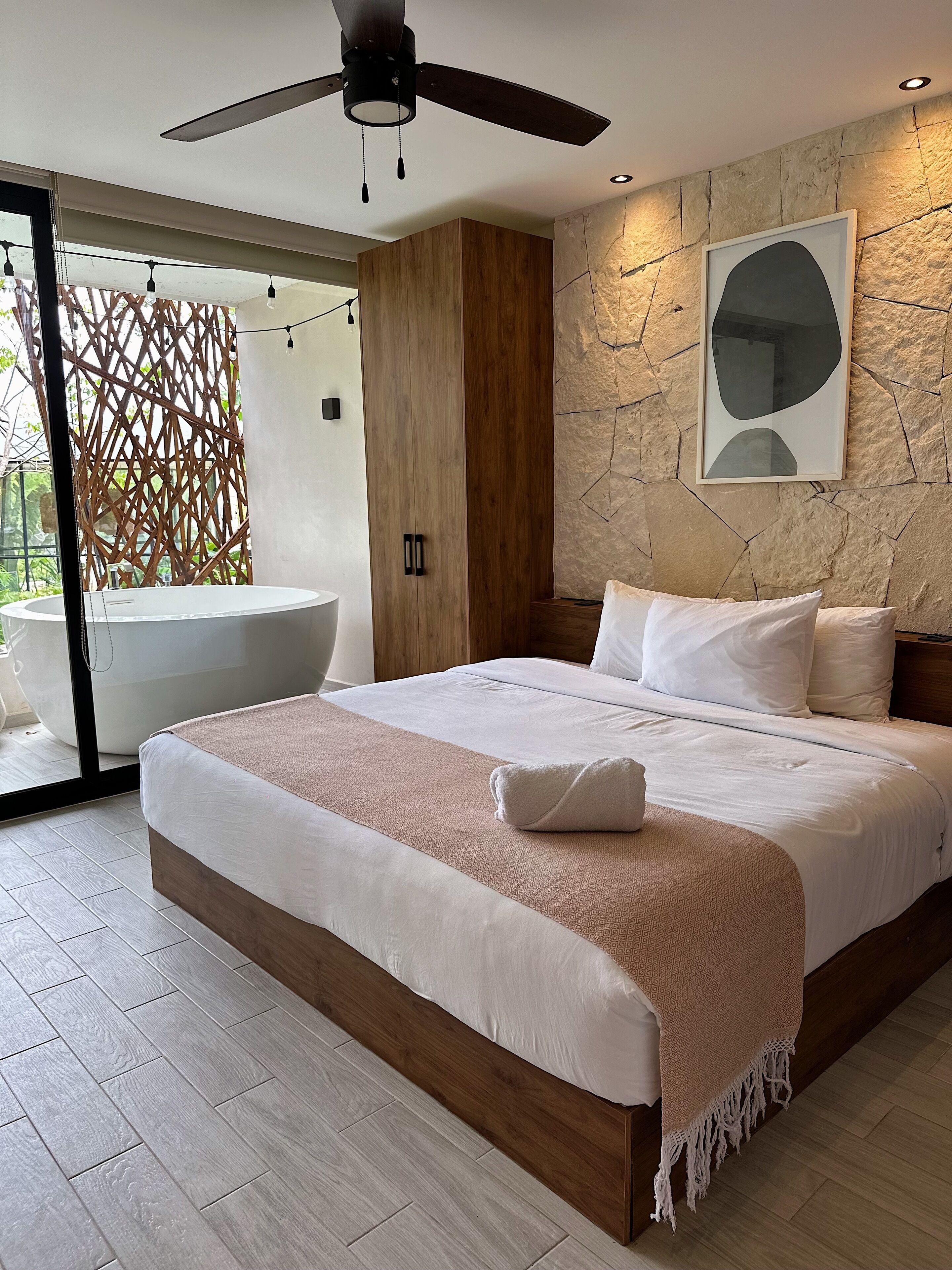 A modern bedroom with a large bed, a ceiling fan, a stone wall, and a bathtub visible through a glass door.