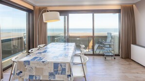 A hotel room with a large table, chairs, a lamp, and a view of the beach through large windows. - Corner Apartment With Frontal sea View (Middelkerke)