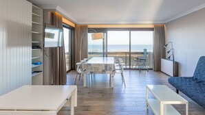 A spacious living area with a balcony overlooking the sea, a dining table with chairs, and a blue sofa. - Corner Apartment With Frontal sea View (Middelkerke)