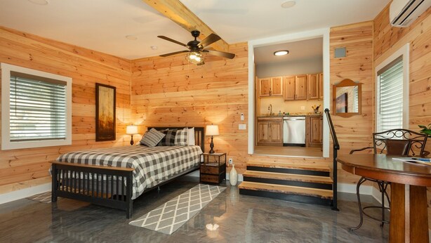 Ein schlafzimmer im Chalet-Stil mit Bett, Nachttischlampen, Deckenventilator und Blick auf die Küche durch einen offenen Durchgang.