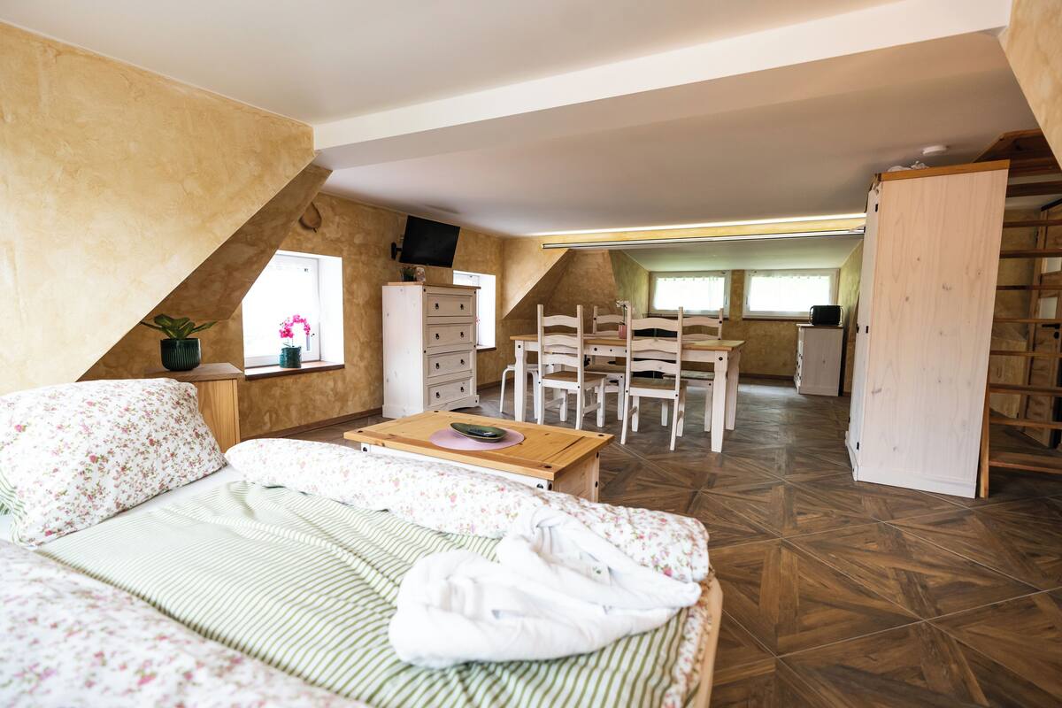 Un dormitorio con cama, escritorio y silla. Se ve una ventana con cortinas, un aparador y un televisor en la pared.