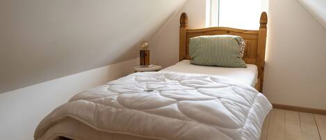 Una cama individual con cabecera de madera en una habitación con techos inclinados y un ventanal pequeño.