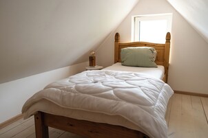 Una cama individual con cabecera de madera en una habitación con techos inclinados y un ventanal pequeño.