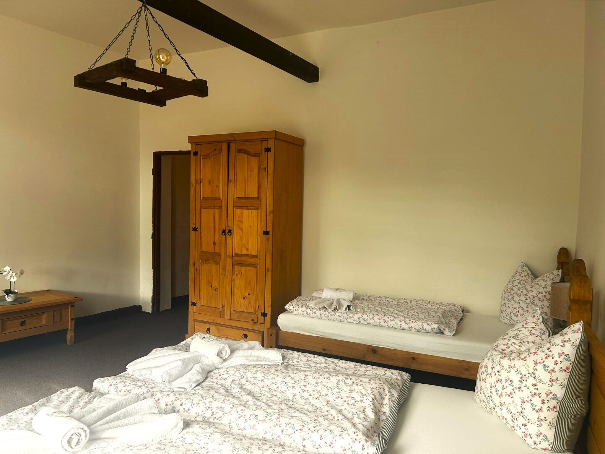 A bedroom with a wooden wardrobe, a bed with floral bedding, a bedside table, and a hanging light fixture.
