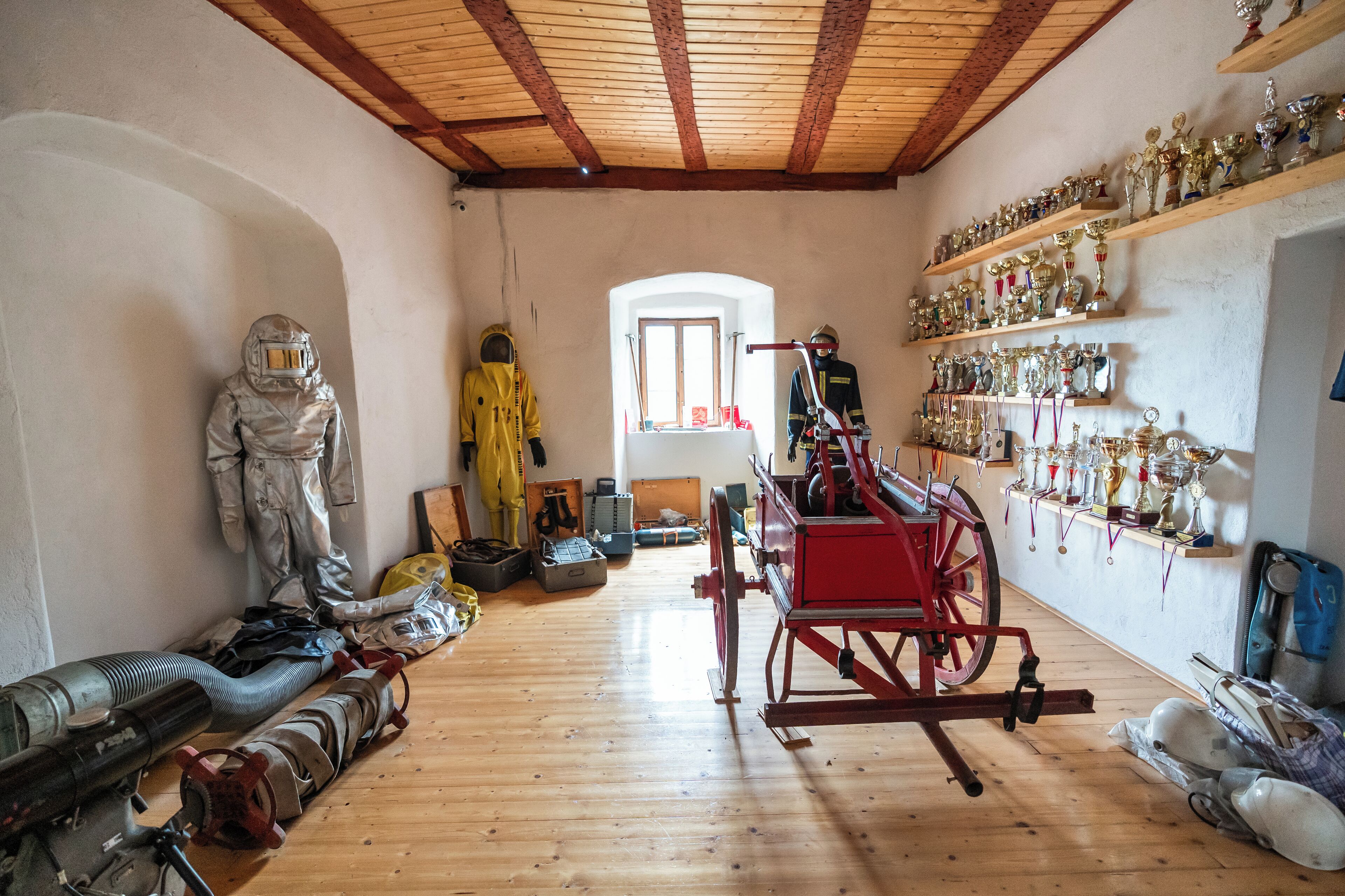 Quarto com piso de madeira, carrinho vermelho, manequins com roupas de proteção e prateleiras com troféus.