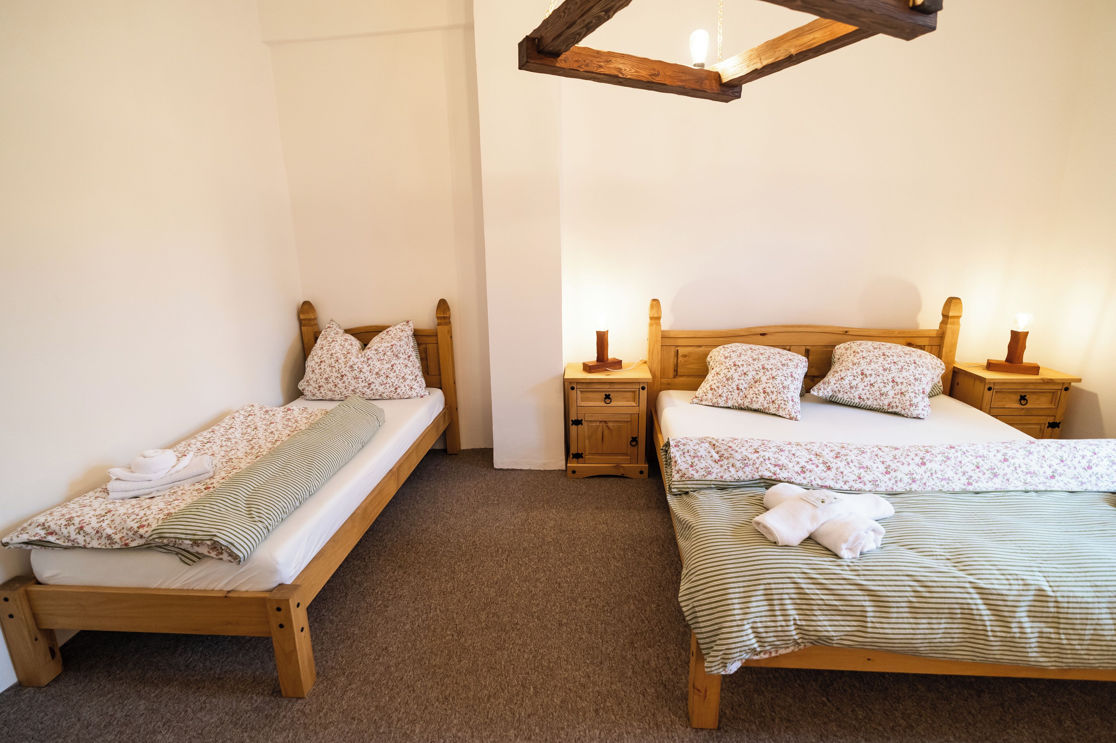 Dos camas de madera con ropa de cama floral, una mesita de noche de madera con lámpara y un sencillo suelo cubierto de alfombra.
