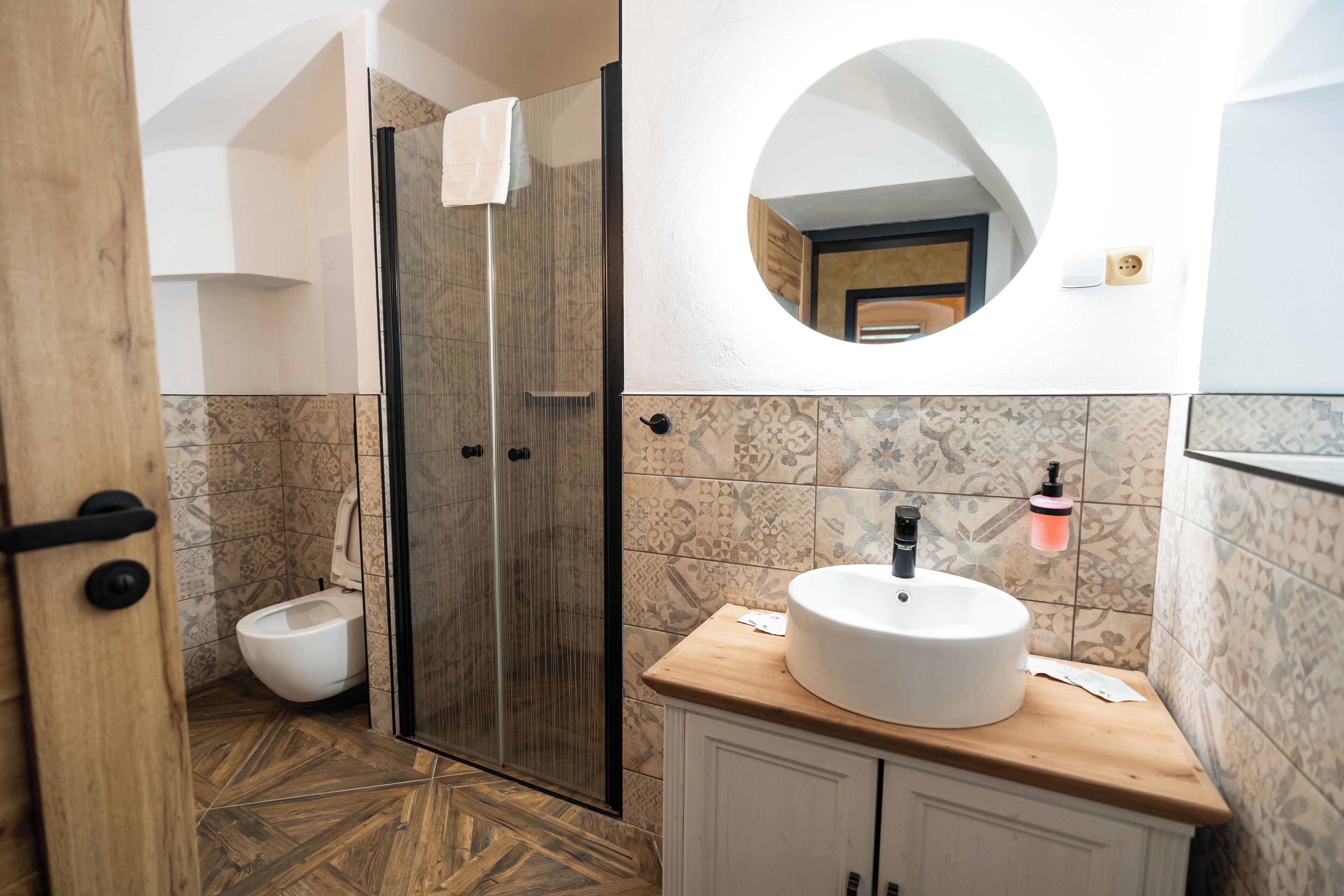 A bathroom with a wooden floor, a white sink, a glass shower enclosure, and patterned tiles.