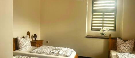 A bedroom with two beds, a wooden headboard, a bedside table with a lamp, a window with blinds, and a small potted plant.