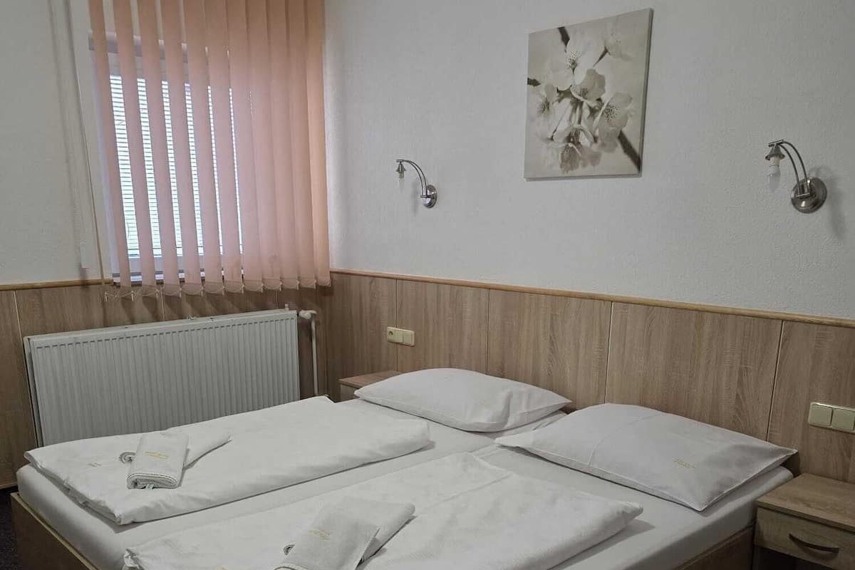 Cama doble con ropa blanca, cabecera de madera, mesita de noche, lámpara de pared y ventana con persianas.