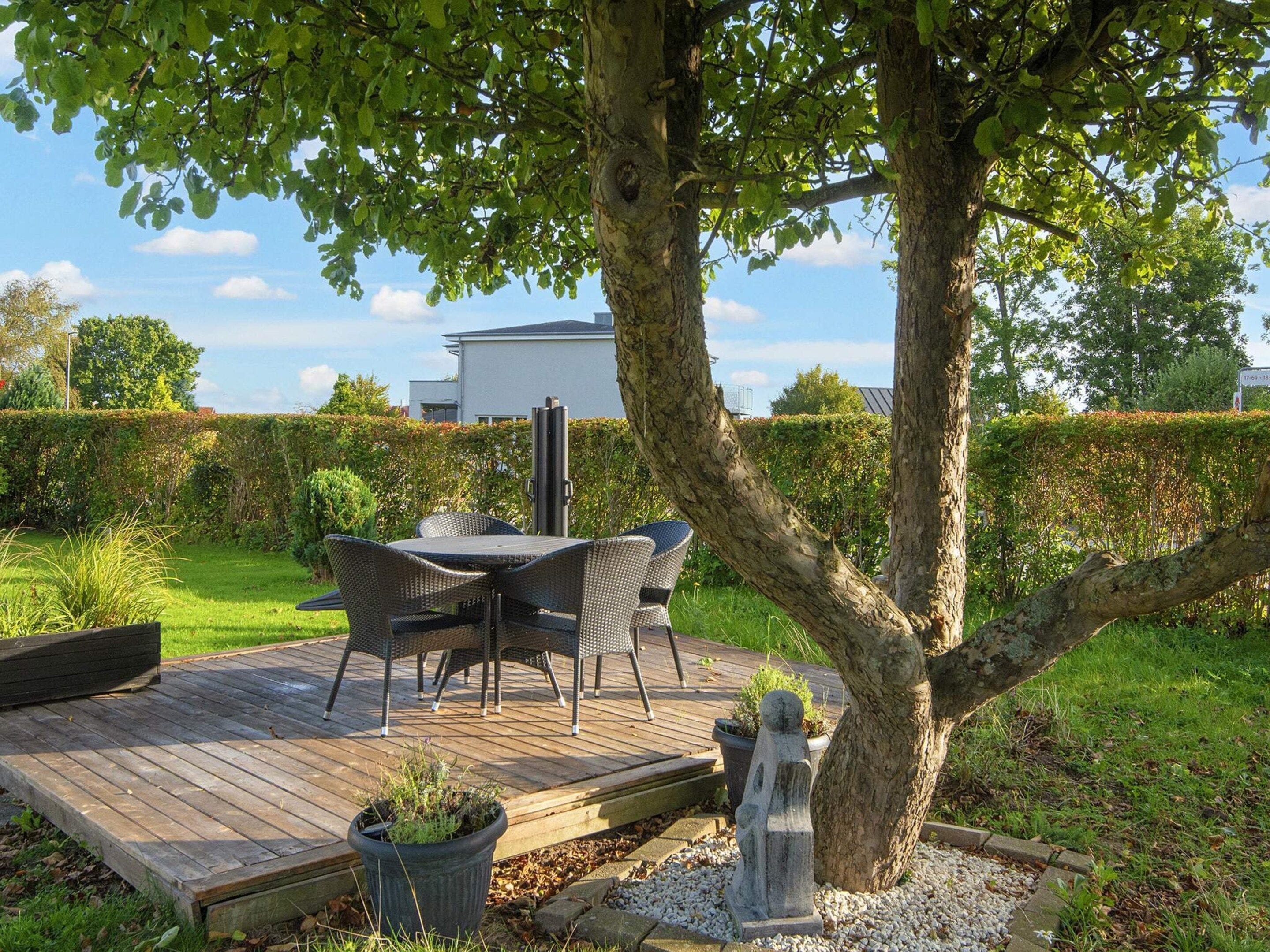An outdoor patio with a table and chairs, surrounded by greenery and a hedge.