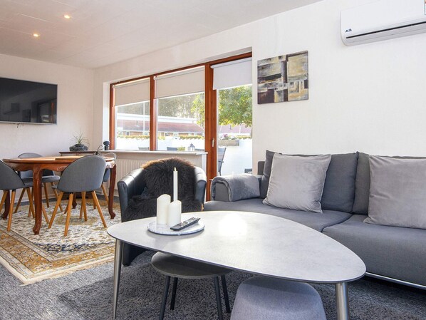 A modern living room with a sofa, a dining table with chairs, a TV mounted on the wall, and a large window with a view of the outside.