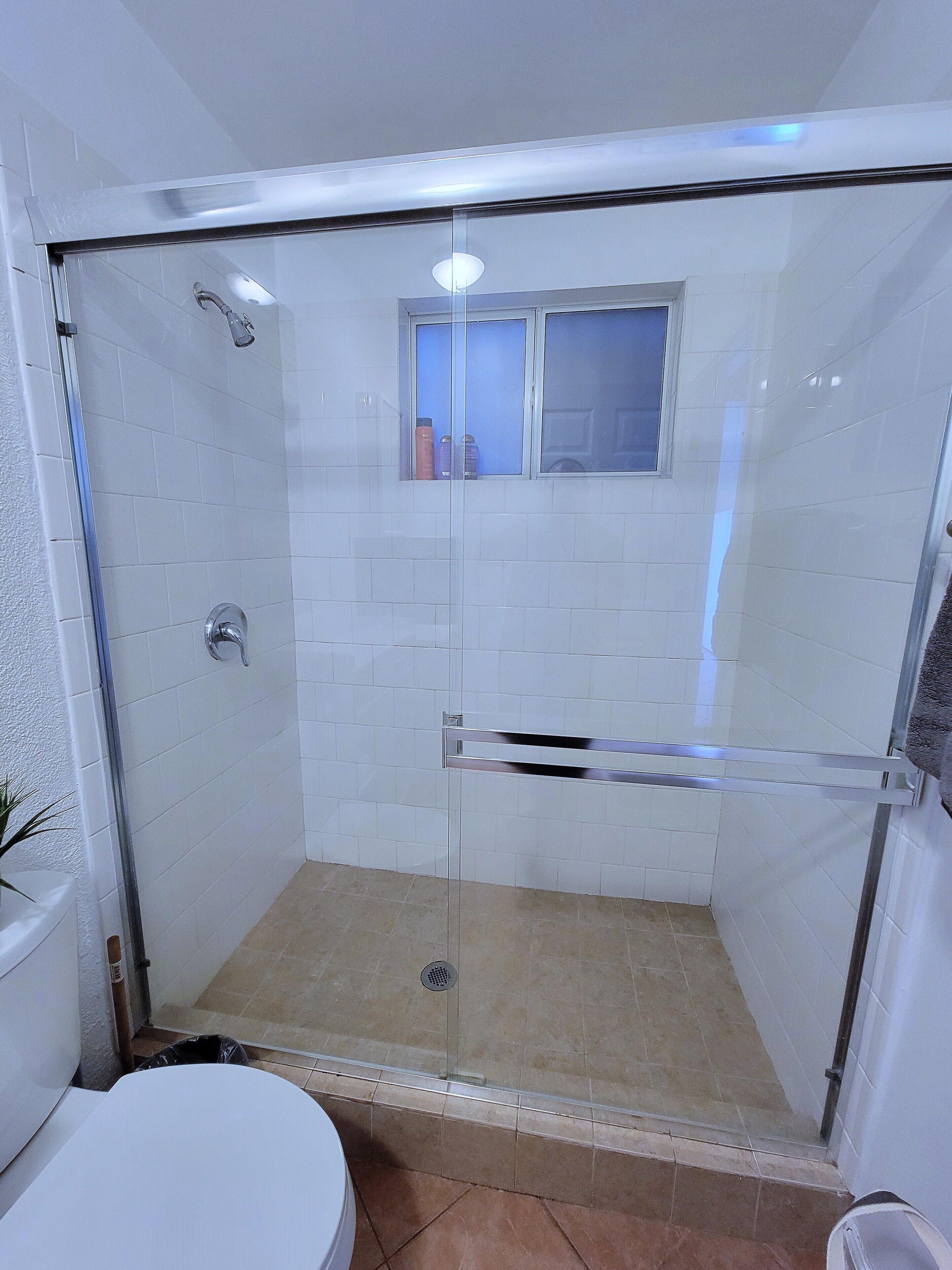 Ducha con cerramiento de vidrio, ventana, paredes de azulejos blancos y desagüe visible.