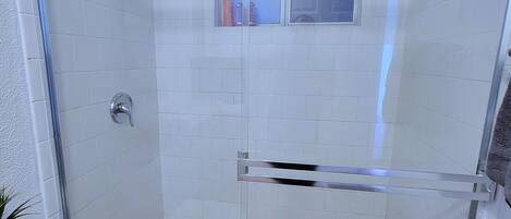 Ducha con cerramiento de vidrio, ventana, paredes de azulejos blancos y desagüe visible.