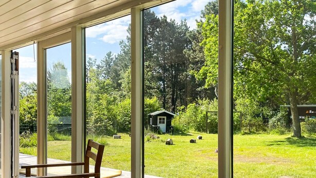 A room with large windows, a wooden deck, and a view of a grassy area with trees and a small building. - 7 Person Holiday Home in Sjaellands Odde (Sjællands Odde)