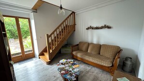 Una sala de estar con una escalera de madera, un sofá, una mesa de centro con revistas y vistas a través de un ventanal.