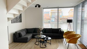 A modern living room with a sectional sofa, a coffee table, and yellow chairs. A staircase is visible on the left, and there is a large window with blinds. - Water Villa in Heeg by Heegermeer (Heeg)