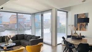 A modern living room with a sectional sofa, a dining table with chairs, and large windows with blinds. - Water Villa in Heeg by Heegermeer (Heeg)