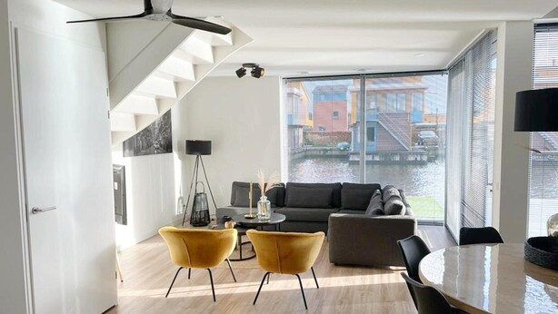 A modern living space with a staircase, a sofa, two yellow chairs, a round table, and a large window with a view of buildings. - Water Villa in Heeg by Heegermeer (Heeg)