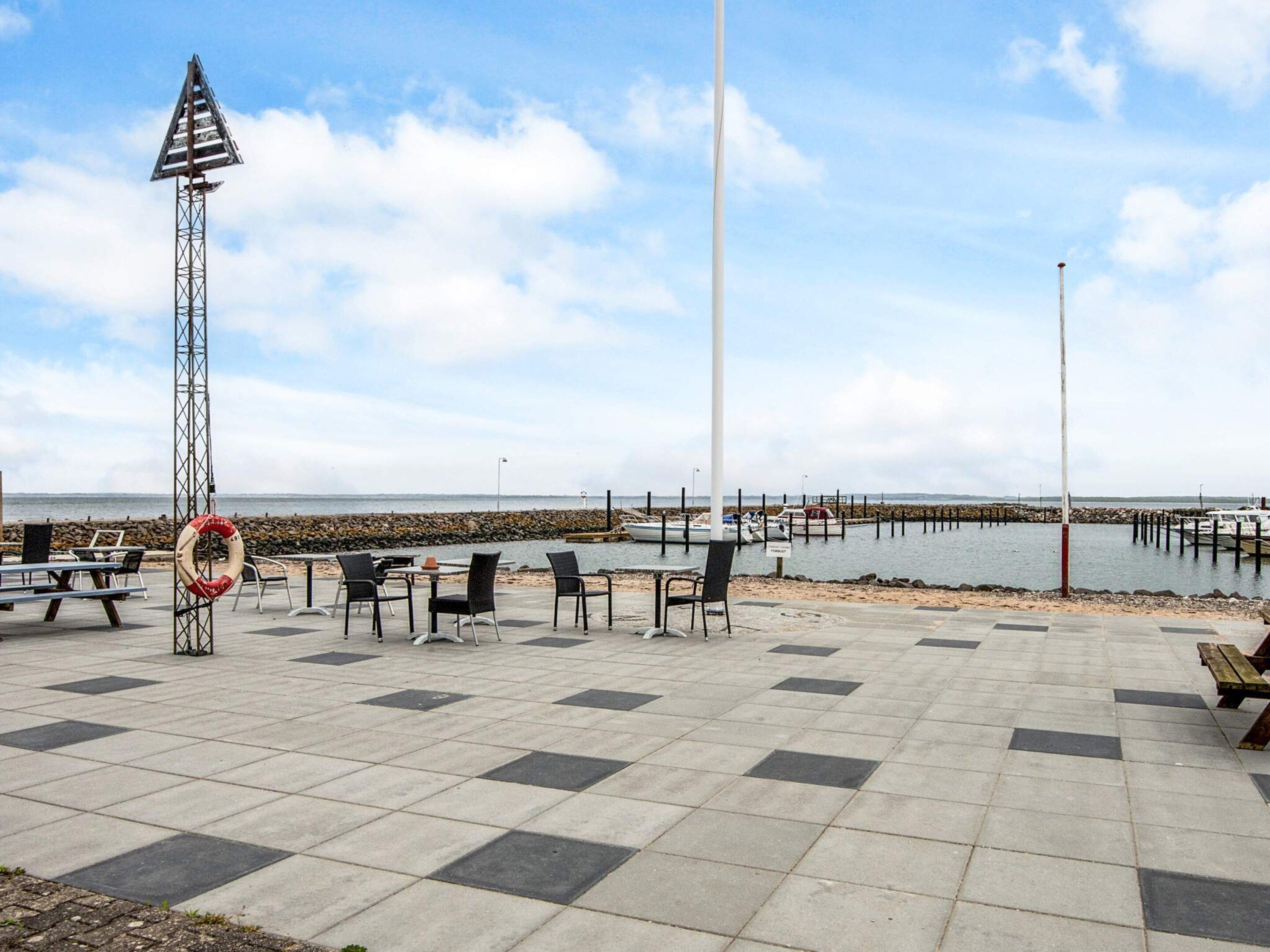 A seaside terrace with tables and chairs, a lifebuoy, and a pier in the background.