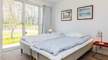 A bedroom with a large window, a bed, a bedside table with a lamp, and two framed pictures on the wall.