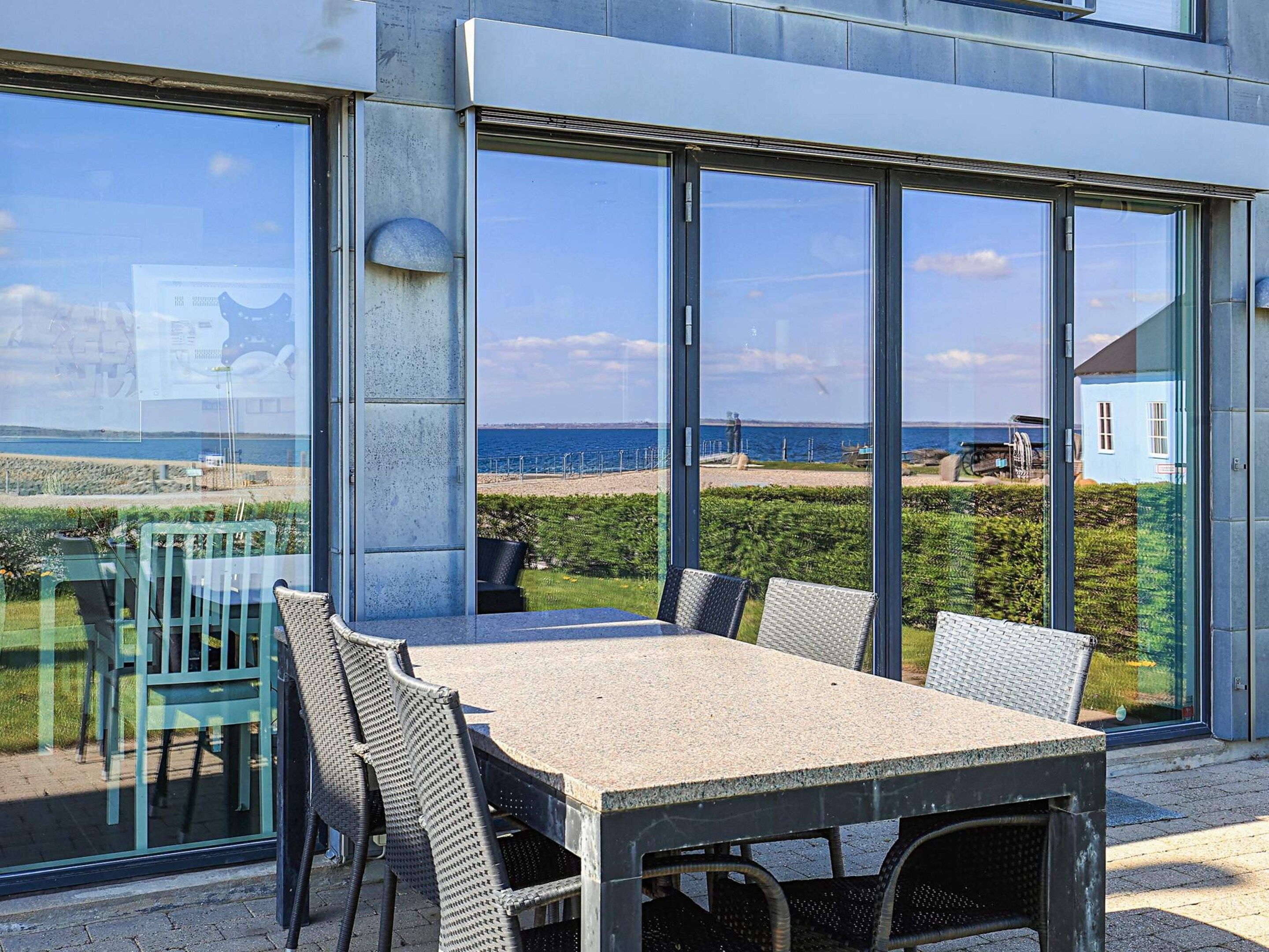 Ein moderner Außenbereich zum Essen mit Tisch, Stühlen, großen Glasfenstern und Blick auf Strand und Meer.