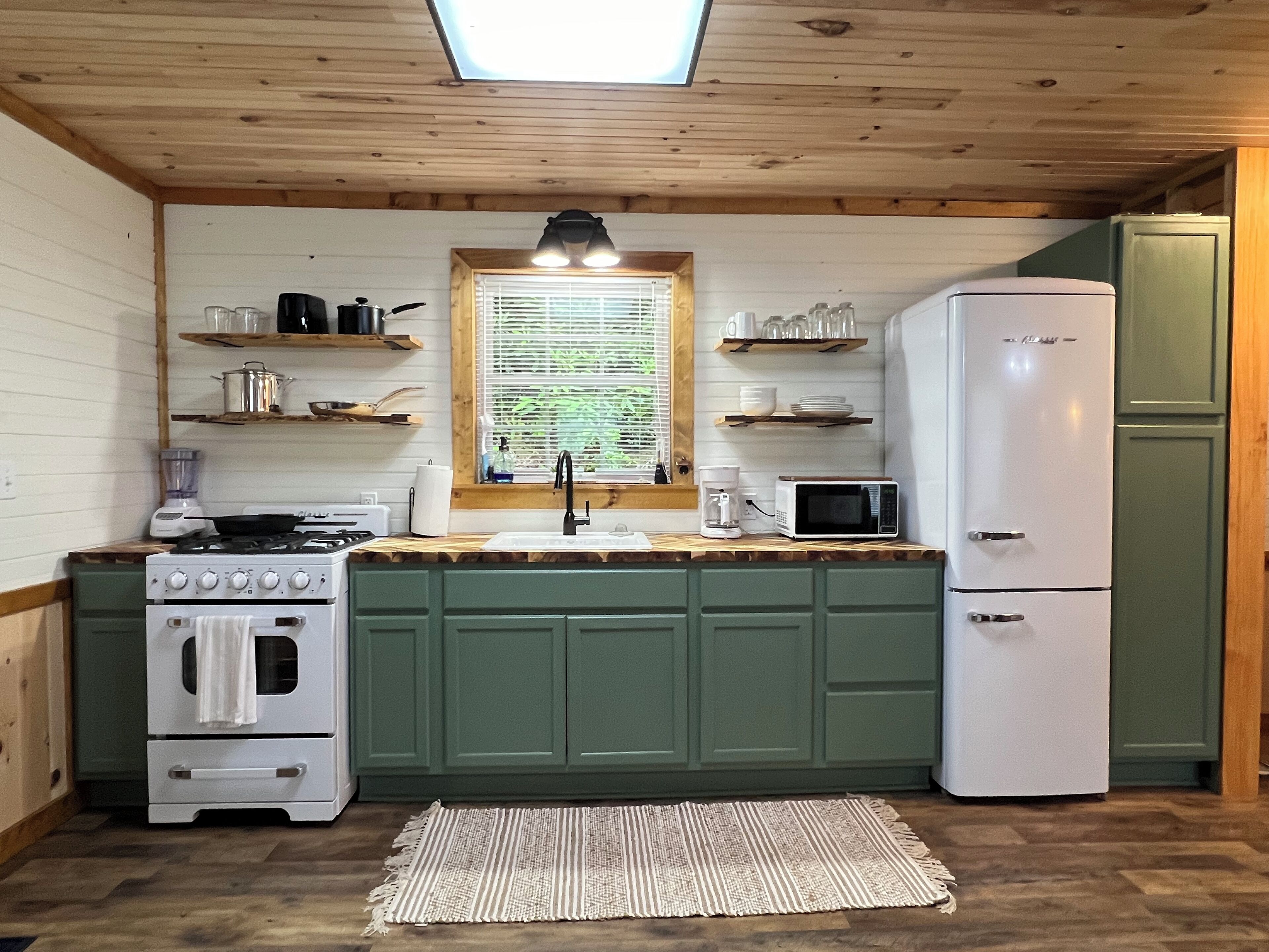 Une cuisine compacte équipée d’une cuisinière à gaz, d’un four à micro-ondes et d’un réfrigérateur. On y trouve des étagères en bois au-dessus de l’évier et une fenêtre avec des stores.