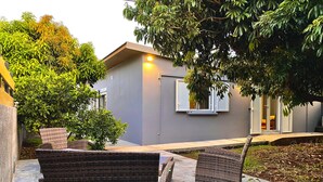 A modern house with a grey exterior, a small patio with wicker furniture, and a tree providing shade. - Ti Case Fleur Letchi - Cadre Calme et Agréable, Avec Jardin (Les Avirons)