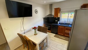 A compact kitchen with a dining table, chairs, a TV mounted on the wall, and a window with a view of greenery. - Ti Case Fleur Letchi - Cadre Calme et Agréable, Avec Jardin (Les Avirons)