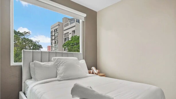 A neatly made bed with white linens and pillows, a window with a view of buildings and trees, and a small bedside table with a rolled towel. - #3 - Charming Spanish Colonial Apartment - Miramar (San Juan,)