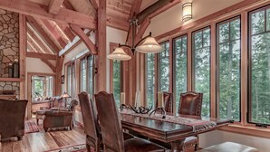 Un comedor rústico con una gran mesa de madera, sillas de cuero y un banco. Se observan amplias ventanas con vistas a un bosque, paredes de piedra y vigas de madera.