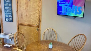 Ein Essbereich mit einem runden Holztisch und vier Stühlen, einer Chandelüster, einem Fernseher, der 'DreamWorks Trolls' zeigt, und einem Schild mit Hausregeln.