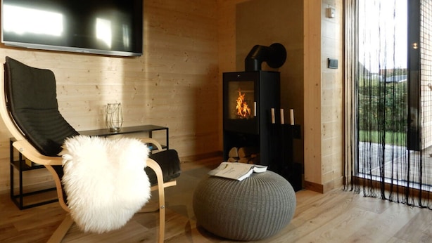 A modern living room with a wooden interior, a black sofa, a glass coffee table, a round ottoman, a fireplace, and a large TV mounted on the wall. - Holiday Apartment in Thuringia (Zierow)