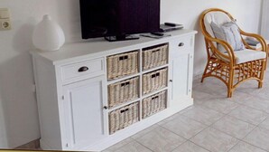 A white cabinet with wicker baskets, a flat-screen TV, and a wicker chair with cushions. - Seebergweg Modern Retreat (Maasholm)
