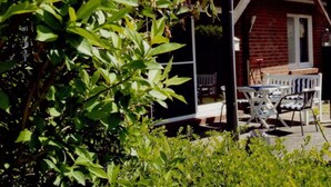 A covered patio with a brick house in the background, a table with chairs, and lush greenery in the foreground. - Seebergweg Modern Retreat (Maasholm)