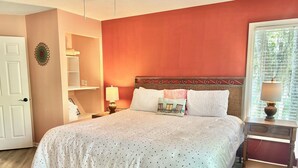 Un dormitorio con una cama grande, una mesita de noche con lámpara, un estante con toallas dobladas y una puerta con picaporte.