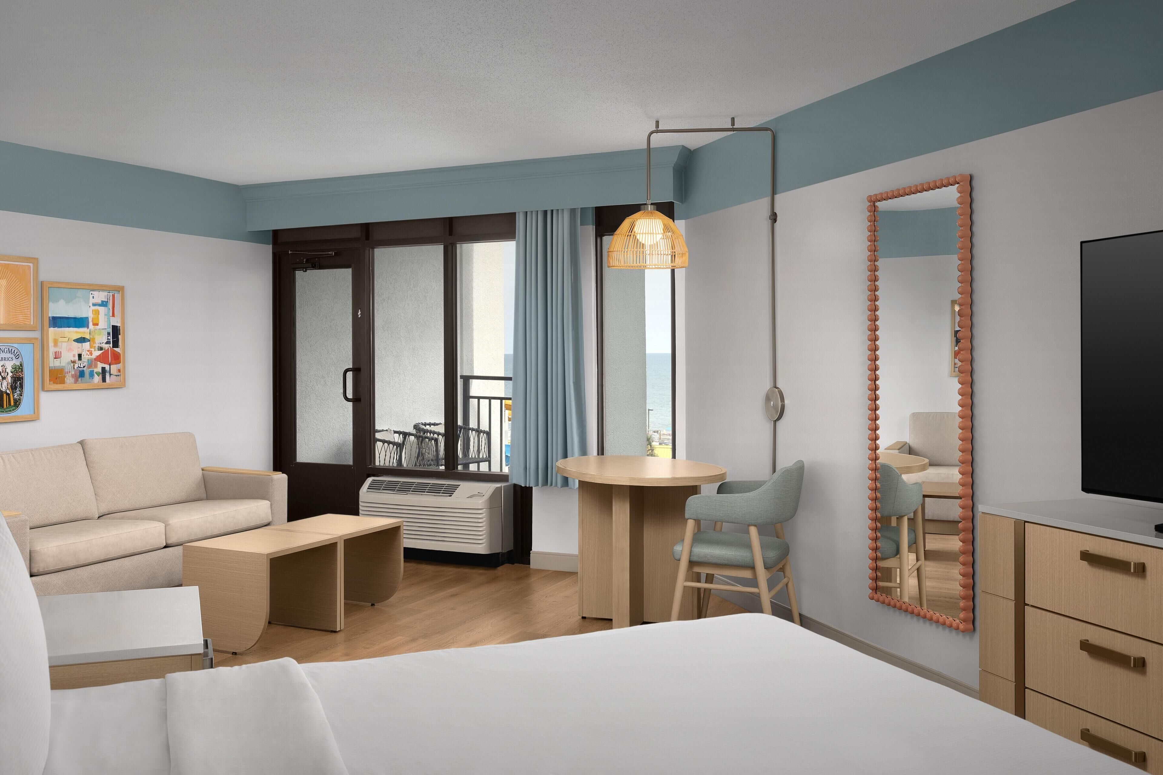 A modern hotel room with a bed, a sofa, a desk, and a chair. There is a large mirror and a television on the wall.