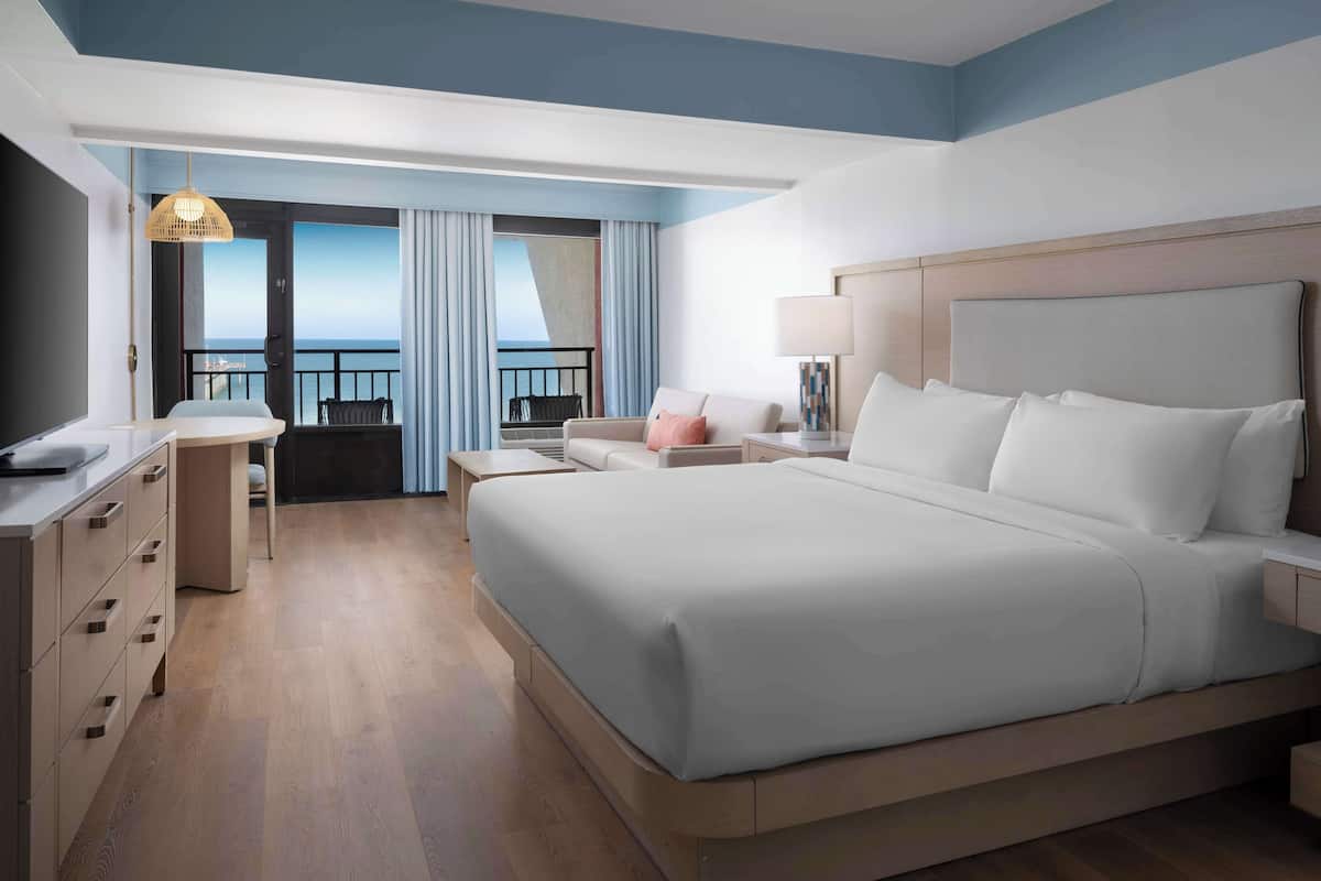 A modern hotel room with a large bed, a desk, and a balcony with a view.
