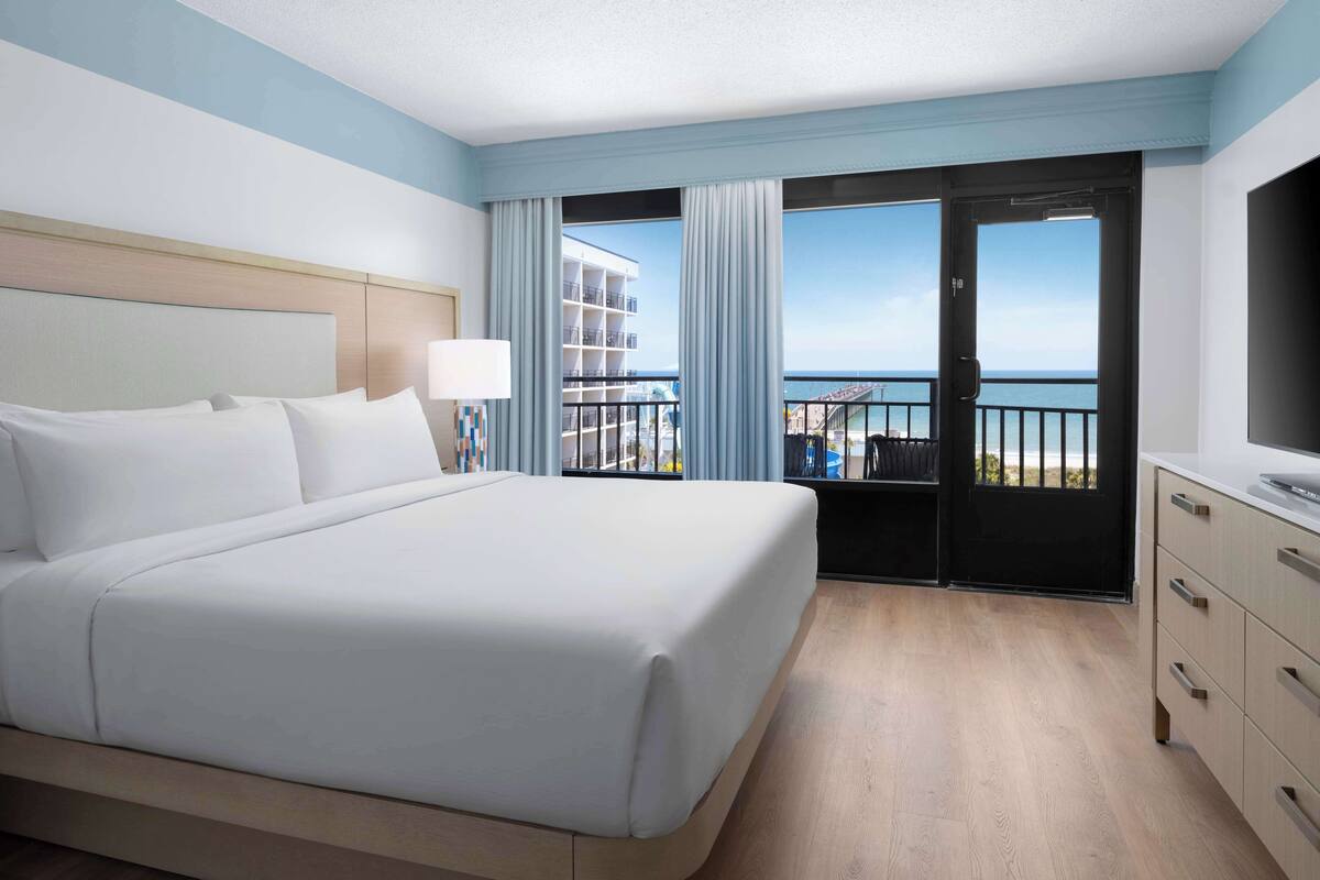 A hotel room with a large bed, a TV, a dresser, and a balcony with a view of the sea and buildings.