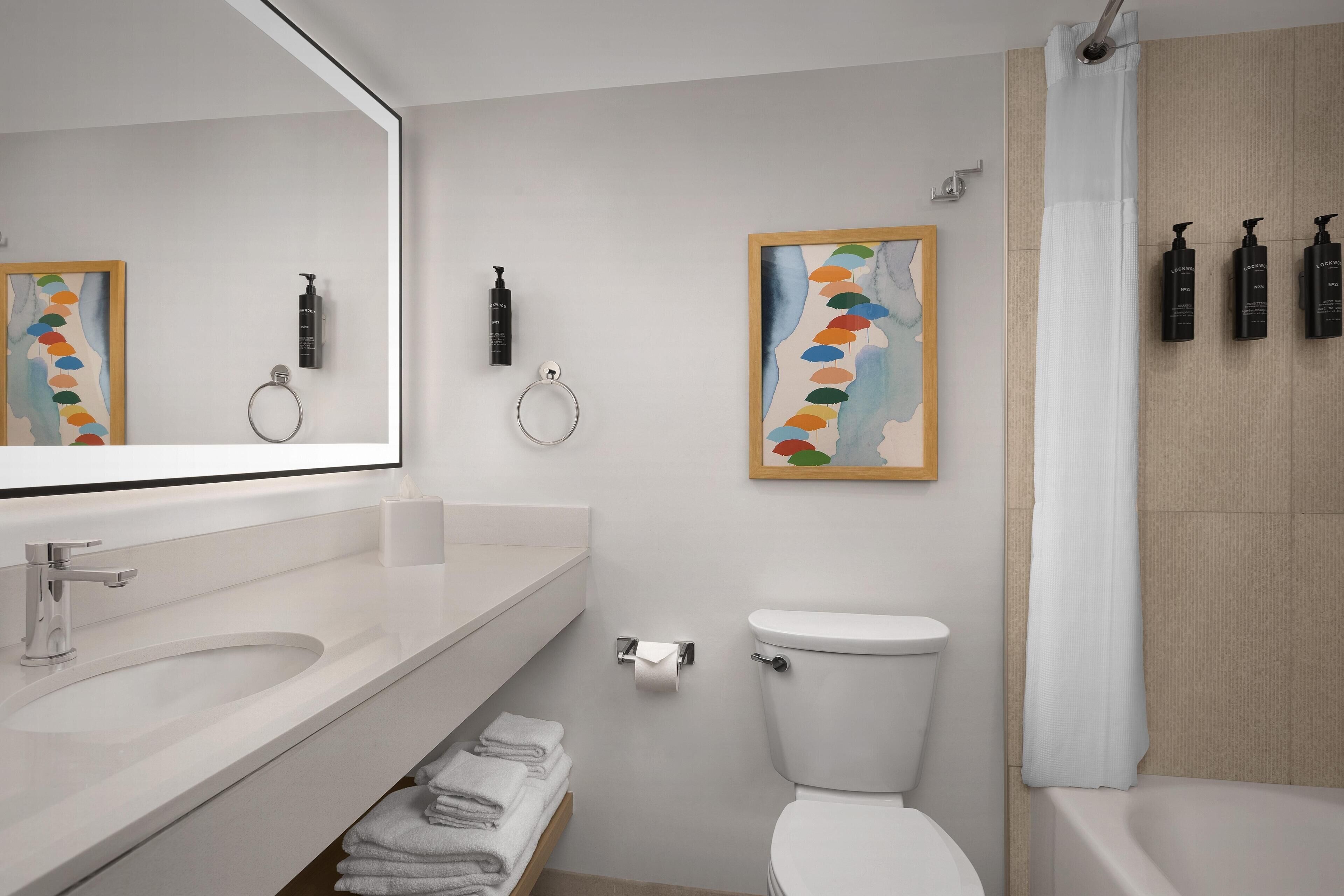 Une salle de bain moderne avec un lavabo, des toilettes et une baignoire, tous de couleur blanche. On y trouve des serviettes pliées sur une étagère, une œuvre d’art encadrée au mur et des articles de toilette accrochés au mur.