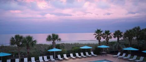 A poolside area with lounge chairs and umbrellas, overlooking a beach and ocean.